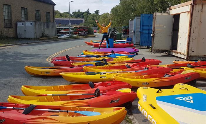 Lots of colourful kayaks and paddleboards ready to be issued to customers with an excited person jumping in the air